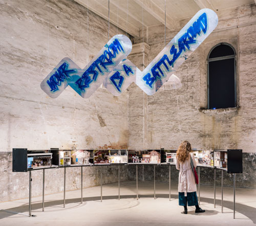 woman standing in room looking at display; text above reads "Your Restroom is a Battleground"